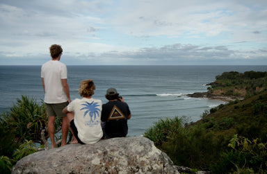 Three people sit on a large rock overlooking a coastal view with waves breaking near the shore.