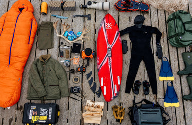 A flat-lay arrangement of essential winter surfing gear on a wooden dock. Includes a bright orange sleeping bag, wetsuit, surfboard, winter clothing, and various camping tools.