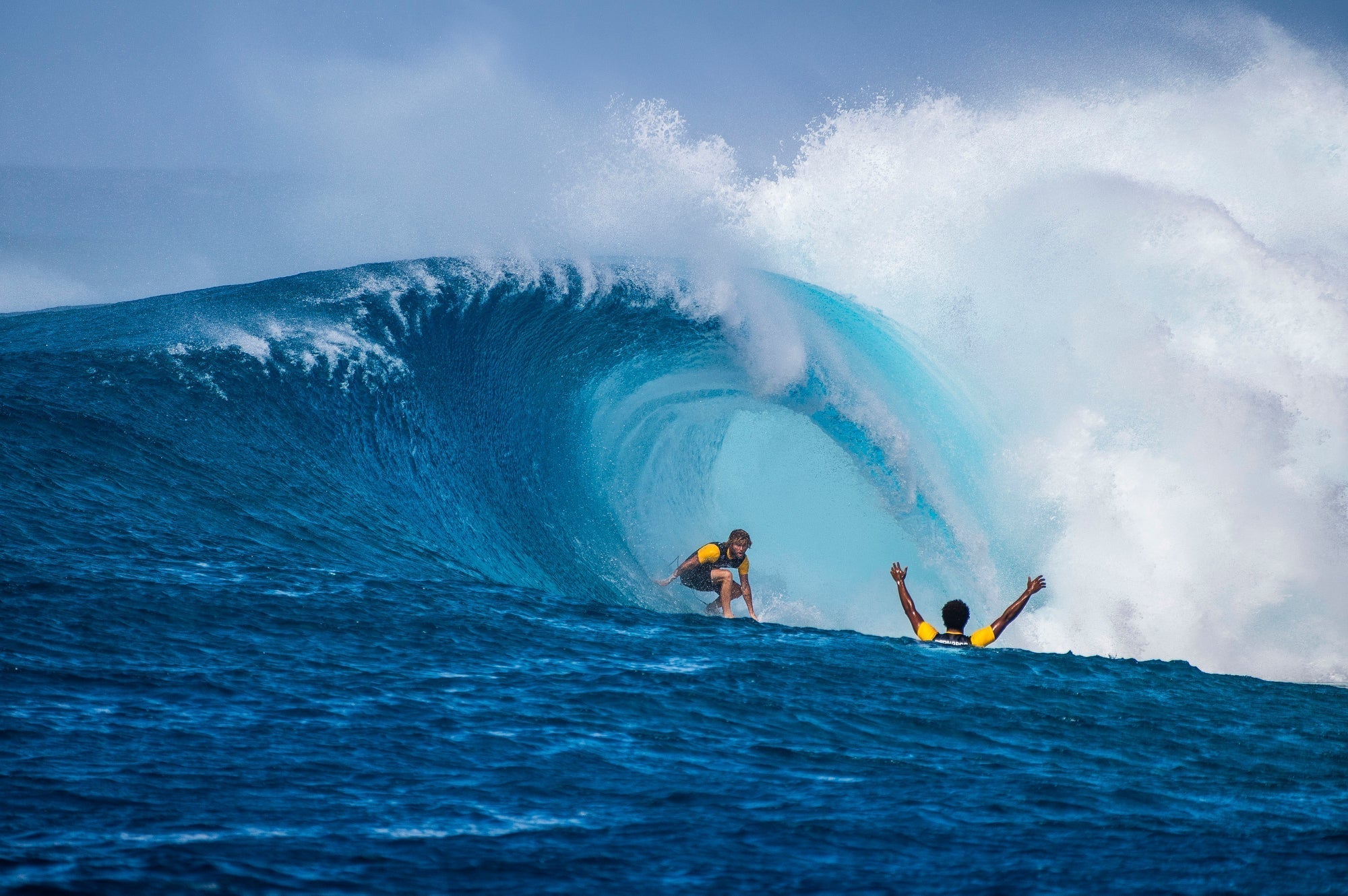 Surfer riding a massive turquoise wave, perfectly positioned inside the barrel, showcasing expert surfing skills in powerful ocean conditions. Another surfer paddles in the foreground, adding depth to the dynamic scene. 