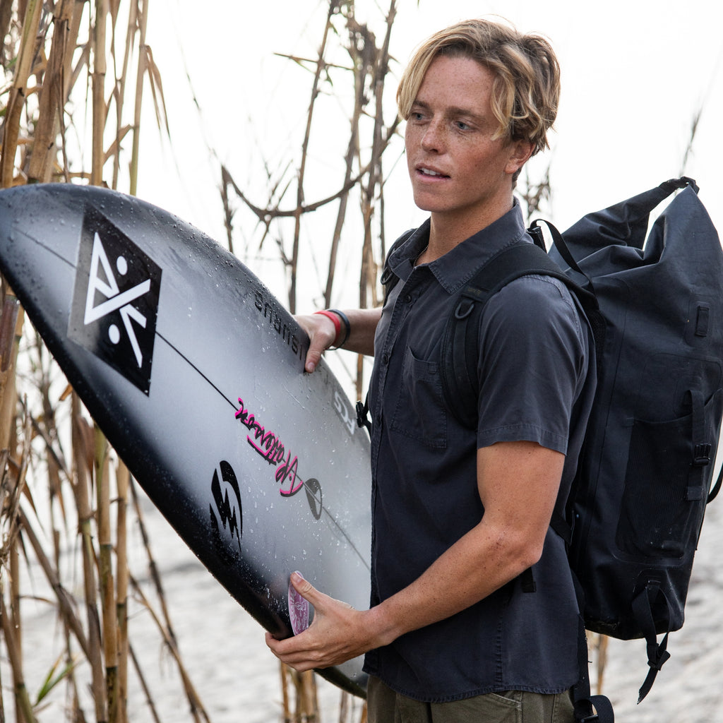 Man holding a surfboard with a Roark logo, standing outdoors near reeds. Big Image - 2