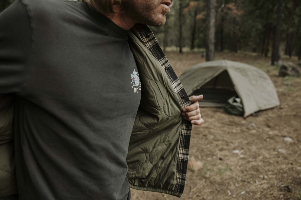 A man wearing a Roark Backwoods shirt in a blue color, standing with his arms crossed in a forest setting.