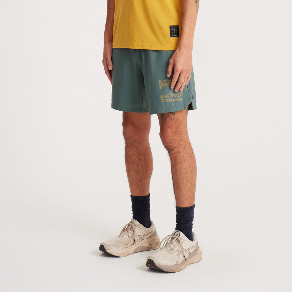 Side angle of a male model wearing green running shorts with 'Open Roads Open Minds' graphic and mustard yellow T-shirt. Big Image - 4