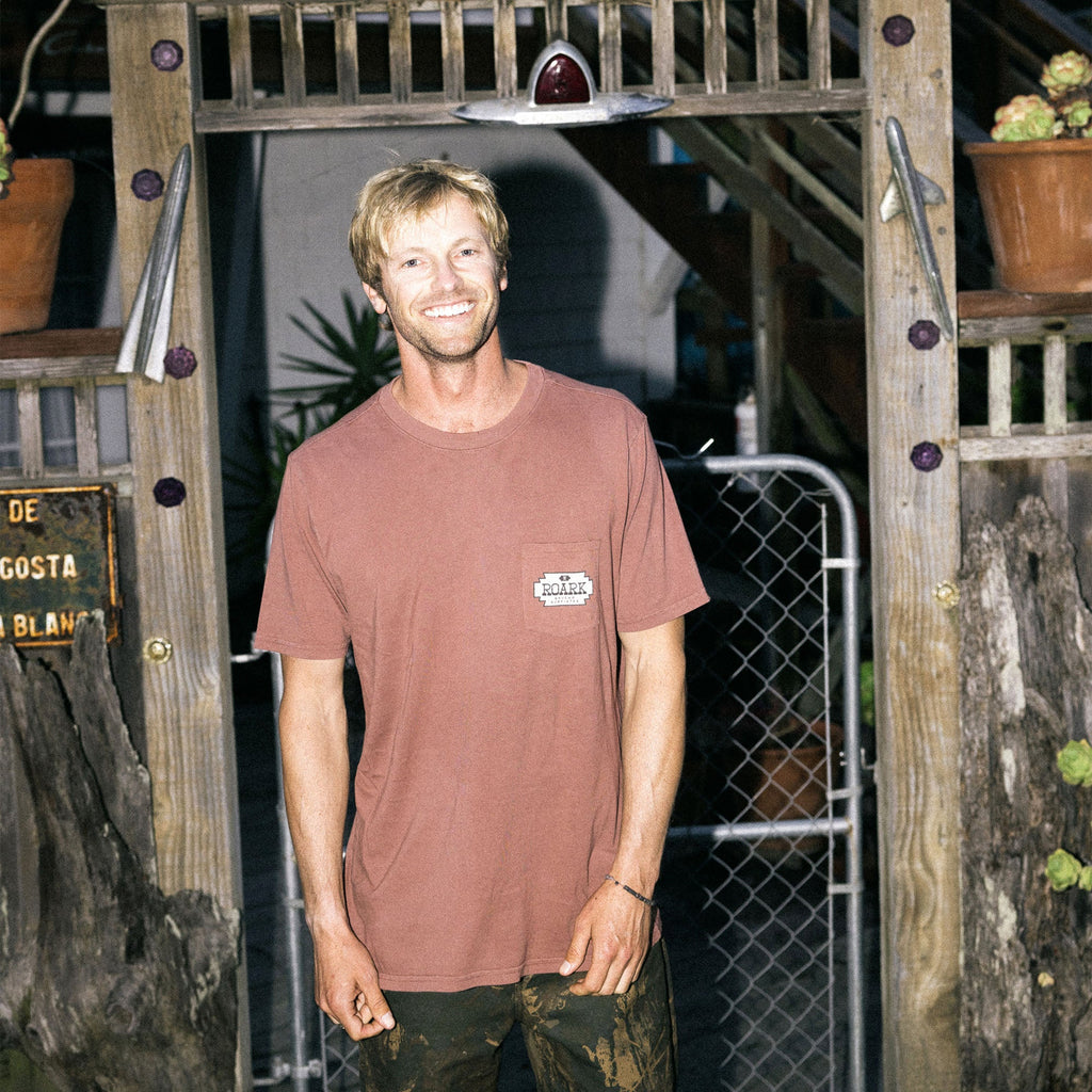 Smiling man with facial hair wearing a Roark Tierra del Sur Shield Pocket Premium Tee, showcasing active outdoor style against a wooden backdrop. Big Image - 2